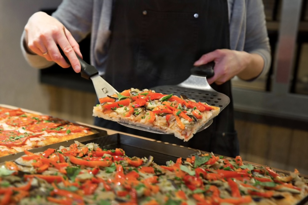 Part de pizza romaine aux légumes colorés tenue à la spatule au-dessus du comptoir